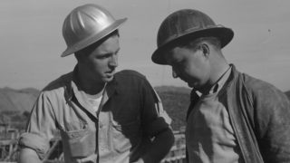 A black-and-white image of two male construction workers, wearing hard hats, in conversation.