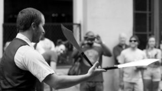 Black-andwhite photograph of Stewart Lawrence Sinclair juggling knives in front of a street audience