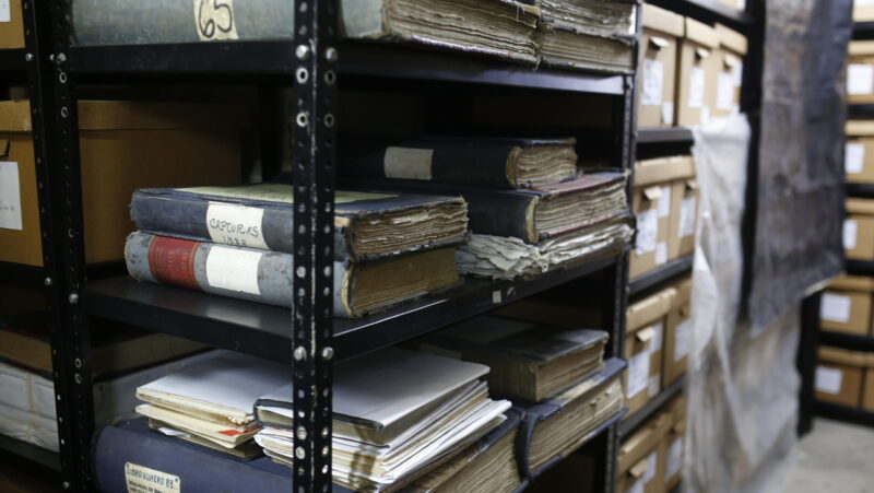 Books and manuscripts stacked on a shelf in an archive, with an aisle of boxes visible in the background